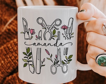 May include: White ceramic mug featuring a black line art design of surgical tools and floral accents. The name "amanda" is written in a script font. The mug is held by a person.