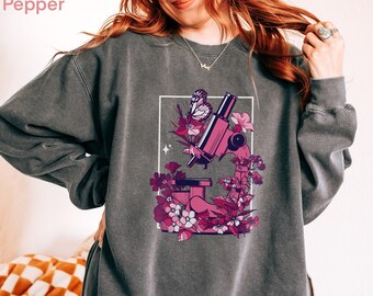 Puede incluir: Sudadera gris oscuro con un diseño de microscopio rosa rodeado de flores rosas y una mariposa. La palabra "Pepper" es visible. La sudadera tiene cuello redondo y mangas largas.