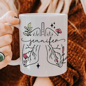 Può includere: Tazza in ceramica bianca con un disegno di polmoni in nero con fiori e vegetazione. Il nome "jennifer" è scritto in corsivo sulla tazza.