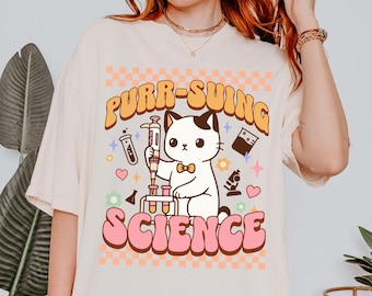 May include: A white oversized t-shirt with a pink and brown graphic of a cat holding a syringe and test tubes. The text "PURR-SUING SCIENCE" is in pink and brown letters.