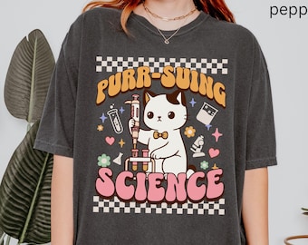 May include: A black t-shirt with a white cat wearing a bow tie and holding a syringe. The text "PURR-SUING SCIENCE" is printed in pink and yellow letters on the shirt.