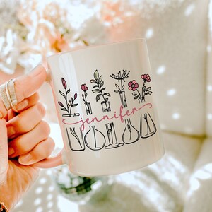 Peut inclure: Une tasse en céramique blanche avec un motif floral et le nom "jennifer" écrit en cursive rose. Le motif représente cinq fleurs dans des fioles en verre avec des tiges et des feuilles.