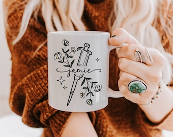 Può includere: Tazza in ceramica bianca con un'illustrazione in bianco e nero di una pipetta e fiori. La tazza ha il nome "Jamie" scritto in corsivo.