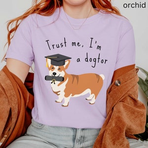 Könnte beinhalten: Ein helles lila T-Shirt mit einem Grafikdruck eines Corgis, der einen Abschluss-Hut trägt und ein Diplom hält. Der Text auf dem Shirt lautet "Trust me, I'm a dogtor."