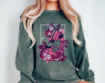 Puede incluir: Una sudadera verde con un microscopio rosa y un diseño floral. El microscopio está rodeado de flores rosas y una mariposa.