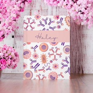 May include: A pink and white notebook with a personalised name, "Haley", on the front. The notebook features a pattern of purple and orange cells, bacteria, and antibodies.