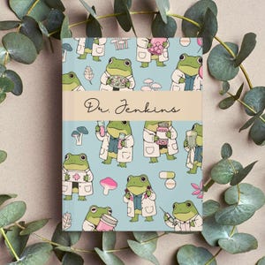 Op de afbeelding: Een notitieboek met een lichtblauwe kaft met een patroon van cartoonkikkers in laboratoriumjassen, die verschillende medische items vasthouden. De tekst "Dr. Jenkins" staat op een beige band.