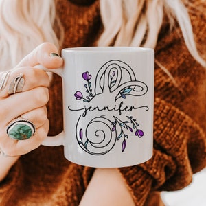 May include: White ceramic mug featuring a floral design with the name "Jennifer" in a script font. The design includes purple flowers and green leaves. The mug is held by a person.
