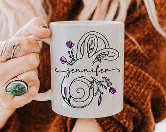 May include: White ceramic mug with a floral design and the name "Jennifer" in a script font. The design includes purple flowers and green leaves. The mug is held by a person.