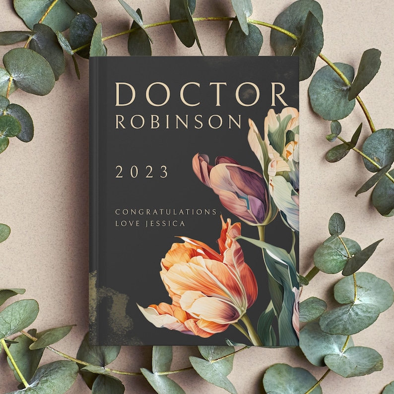 Pu&ograve; includere: Un biglietto nero con lettere dorate che recitano "Doctor Robinson 2023 Congratulazioni Love Jessica". Il biglietto &egrave; decorato con un motivo floreale di tulipani arancioni, viola e bianchi.