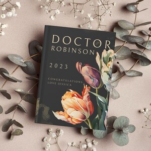 Pu&ograve; includere: Un libro nero con lettere dorate che recitano "Doctor Robinson 2023 Congratulazioni Love Jessica". Il libro &egrave; decorato con un'illustrazione ad acquerello di tulipani arancioni e viola.