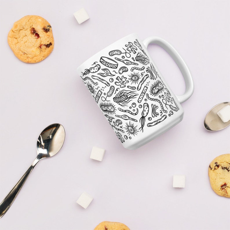 May include: White ceramic mug with a black and white illustration of various bacteria and microbes. The mug is surrounded by sugar cubes, cookies, and a silver spoon.