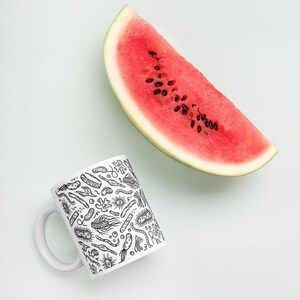May include: A white ceramic mug with a black and white illustration of various bacteria and microorganisms. The mug is next to a slice of watermelon on a light blue background.