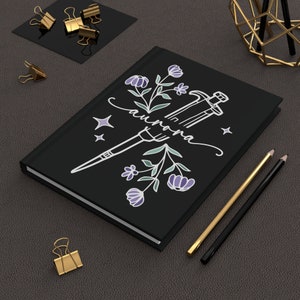 May include: Black notebook with a white illustration of a pipette, flowers, and the word "aurana".