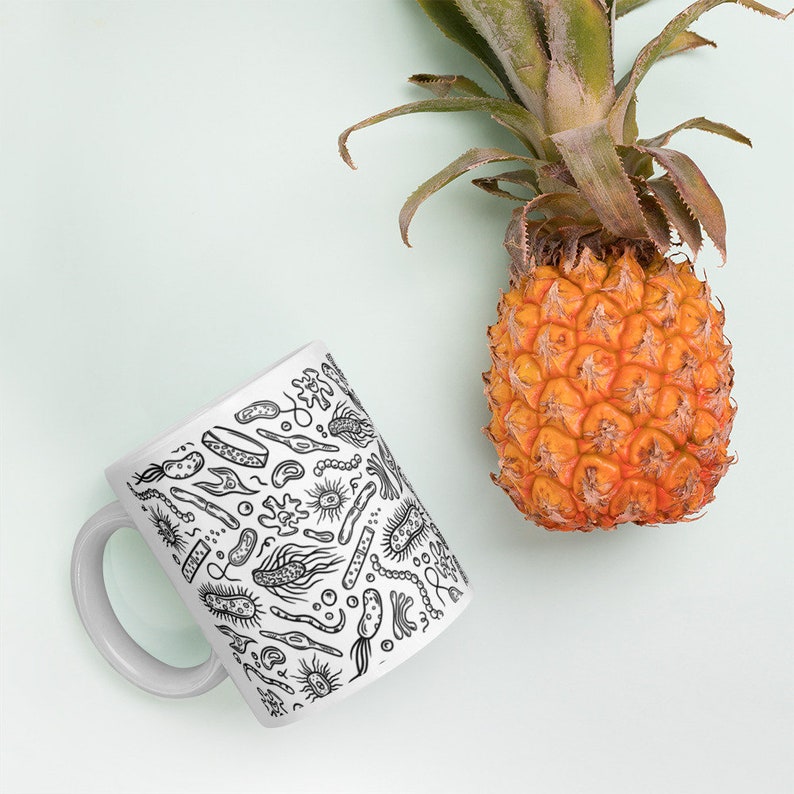 May include: White ceramic mug with a black and white illustration of various bacteria and microorganisms. The mug is sitting on a light green surface next to a pineapple.