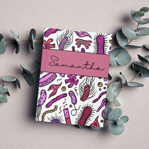 Puede incluir: Una libreta rosa y blanca con un patrón colorido de bacterias y el nombre "Samantha" en la portada.