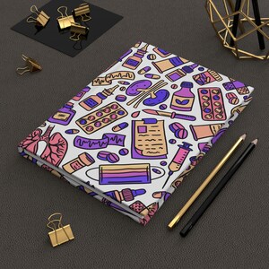 Puede incluir: Un cuaderno blanco con un patrón de garabatos coloridos de suministros médicos, que incluyen píldoras, jeringas, termómetros y un estetoscopio. El cuaderno está sobre una superficie gris oscuro con dos lápices y un soporte de metal dorado.
