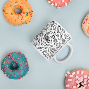 May include: A white ceramic mug with a black and white illustration of various bacteria and microbes. The mug has a handle and is surrounded by colorful donuts.