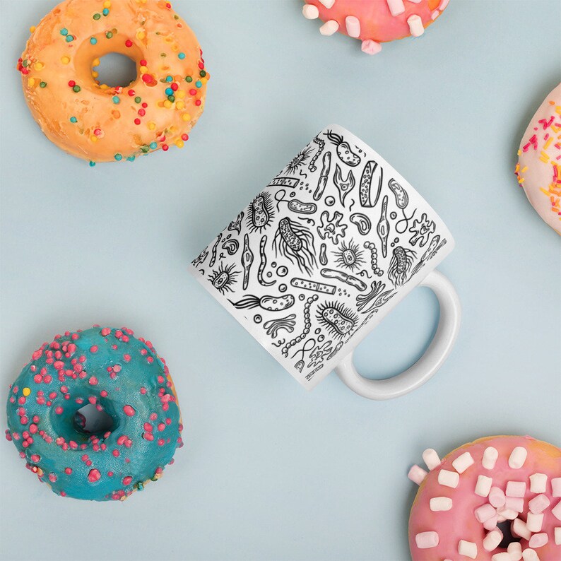 May include: A white ceramic mug with a black and white illustration of various bacteria and microbes. The mug has a handle and is surrounded by colorful donuts.