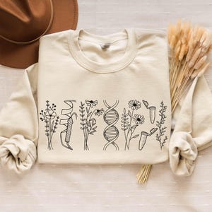 Könnte beinhalten: Ein cremefarbener Pullover mit einem schwarzen grafischen Design aus Blumen, DNA-Strängen und Reagenzgläsern. Der Pullover liegt flach auf einer hellbraunen Oberfläche mit einem braunen Hut und getrocknetem Gras.