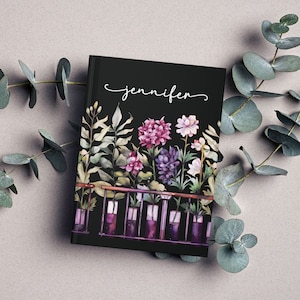 May include: Black notebook featuring a floral design and the name "jennifer" written in white script. The flowers are in various shades of pink, purple, and white and are arranged in a row along the bottom of the notebook.