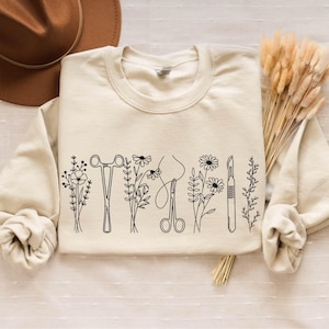 Könnte beinhalten: Ein cremefarbener Pullover mit einer schwarzen Linienzeichnung von chirurgischen Instrumenten und Blumen. Der Pullover liegt flach auf einer hellbraunen Oberfläche.