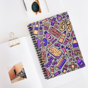May include: A spiral bound notebook with a gray cover featuring a colorful pattern of medical illustrations. The illustrations include pills, syringes, bandages, and medical equipment. The pattern is repeated throughout the cover.