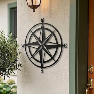 May include: A black metal compass rose wall decor is mounted on a beige wall, with a vintage-style outdoor lamp above it. The compass rose has directional indicators. Potted plants and a wooden door are also visible.