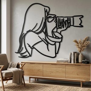 Pode incluir: Uma peça de arte de parede em metal preto com um desenho de linha contínua de uma pessoa com cabelo comprido segurando uma câmara com uma lente grande junto ao olho. O design é minimalista e mostra o sujeito de perfil.