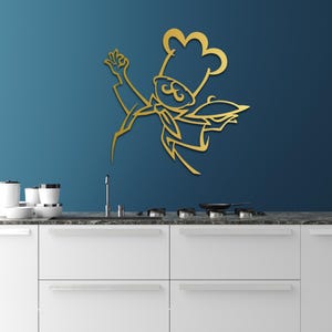 May include: A gold-colored metal wall art piece shaped as a stylized chef wearing a tall toque hat, a neckerchief, and holding a serving platter. The design features clean, sharp lines and a flat silhouette, mounted on a solid dark blue wall.