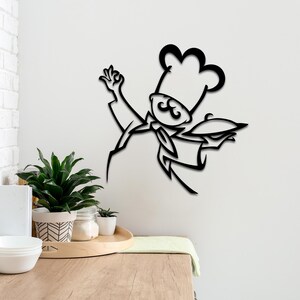 May include: A black metal wall art piece depicting a stylized chef wearing a tall toque hat, a neckerchief, and holding a serving platter with a lid. The design features clean, bold lines and a flat silhouette, mounted on a plain white wall.