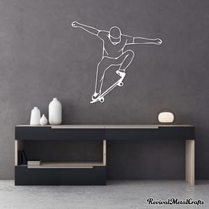 May include: A white metal wall art sculpture of a person skateboarding. The sculpture is mounted on a gray wall above a black shelf with two white vases.