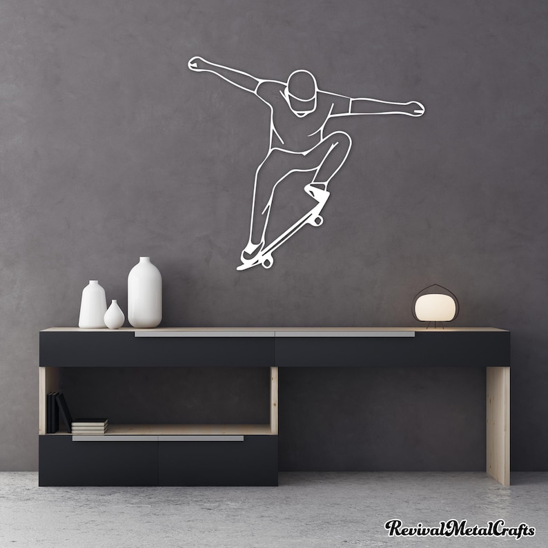 May include: A white metal wall art sculpture of a person skateboarding. The sculpture is mounted on a gray wall above a black shelf with two white vases.