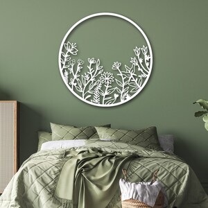May include: White metal wall decor featuring a circular design with a floral pattern. The design is reminiscent of a garden with various flowers and leaves. The decor is mounted on a sage green wall above a green bed with white bedding.