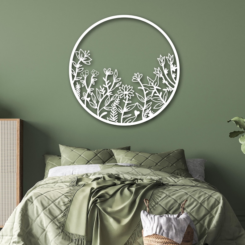 May include: White metal wall decor featuring a circular design with a floral pattern. The design is reminiscent of a garden with various flowers and leaves. The decor is mounted on a sage green wall above a green bed with white bedding.