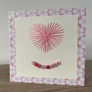 Può includere: Biglietto di auguri fatto a mano con un cuore ricamato rosa. Il biglietto presenta un centro color crema con le parole "with love" scritte con perline colorate. Il biglietto è incorniciato con un motivo floreale rosa.