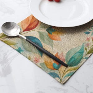 Pode incluir: Uma colher prateada com cabo preto fosco repousa sobre um lugar americano retangular com um padrão estampado de folhas laranja, verde-azulado e verdes e pequenas flores cor-de-rosa. Um prato de cerâmica branca está posicionado sobre o lugar.
