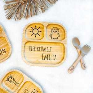 Può includere: Piatto diviso in bambù con un'illustrazione di sole e pinguino e il nome "Emilia". Include cucchiaio e forchetta con "Emilia" incisi sui manici. Il piatto è marrone chiaro con illustrazioni nere.