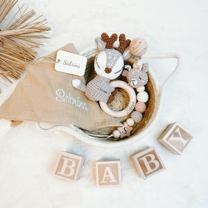May include: A brown crocheted deer toy, a wooden ring, a wooden bead necklace, and a beige muslin burp cloth with the name "Sabrina" embroidered on it, all in a woven basket.  There are also wooden blocks that spell out "BABY" in front of the basket.
