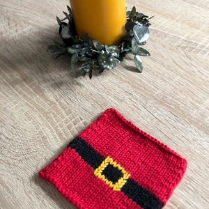 May include: A red knitted coaster with a black stripe and yellow buckle design, reminiscent of Santa's belt. A yellow candle sits on a green wreath. The items are on a wooden surface, creating a festive theme.