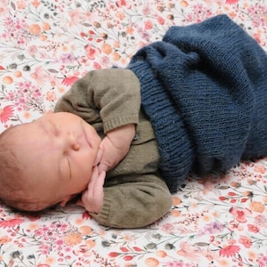 Puede incluir: Un bebé recién nacido durmiendo en una manta con estampado floral. El bebé lleva una manta de punto azul y un suéter de punto verde.