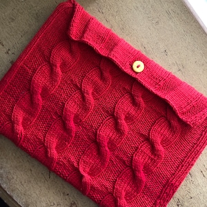 May include: Red knitted laptop sleeve with a cable knit pattern and a yellow button closure.