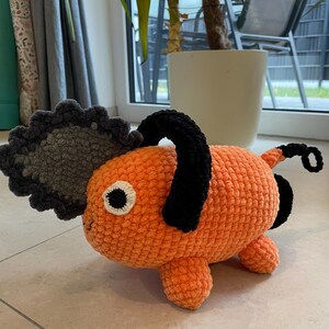 May include: An orange crochet plush toy shaped like a dog with a chainsaw head. The toy has black accents, a gray chainsaw blade, and a single black eye. The plush is sitting on a tiled floor.