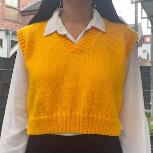 May include: A bright yellow, knitted V-neck vest. The vest is worn over a white collared shirt. The vest has a ribbed hem and a V-neck design. The overall style is casual and preppy.