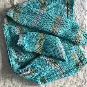 May include: A turquoise and tan knit sweater with long sleeves. The sweater is made of a soft, textured yarn and has a ribbed cuff and hem. The color pattern is a mix of turquoise, tan, and white.