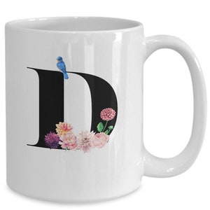 May include: White ceramic mug featuring a large black letter "D" adorned with watercolor flowers in shades of pink, purple, and yellow. A small blue bird perches on the letter. The mug has a curved handle.