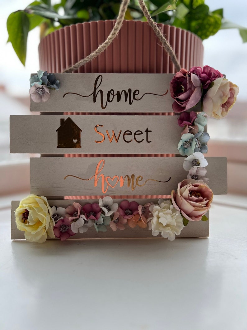May include: A wooden sign with the words "home sweet home" in a cursive font. The sign is decorated with colorful artificial flowers and a jute rope for hanging. The sign has a light beige background.