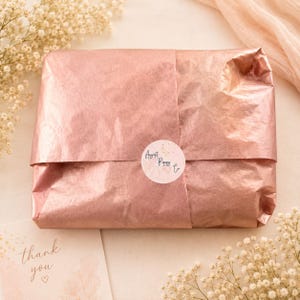 May include: A package wrapped in rose-colored tissue paper, sealed with a round sticker that reads "August Poppy Co." A small card with the words "thank you" is visible. The scene is decorated with sprigs of white flowers.