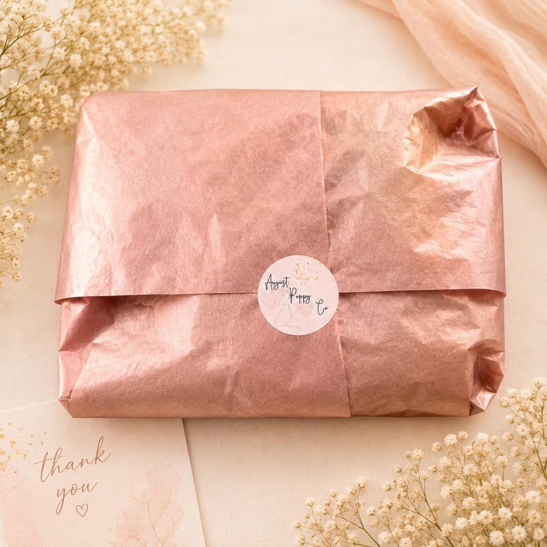 May include: A package wrapped in rose-colored tissue paper, sealed with a round sticker that reads "August Poppy Co." A small card with the words "thank you" is visible. The scene is decorated with sprigs of white flowers.