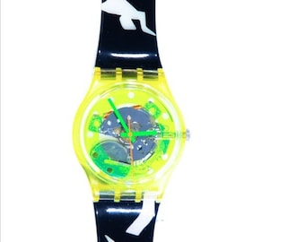 Könnte beinhalten: Eine gelbe Swatch-Uhr mit einem schwarz-weiß gemusterten Armband. Das Zifferblatt der Uhr ist transparent und zeigt das Innenleben der Uhr.
